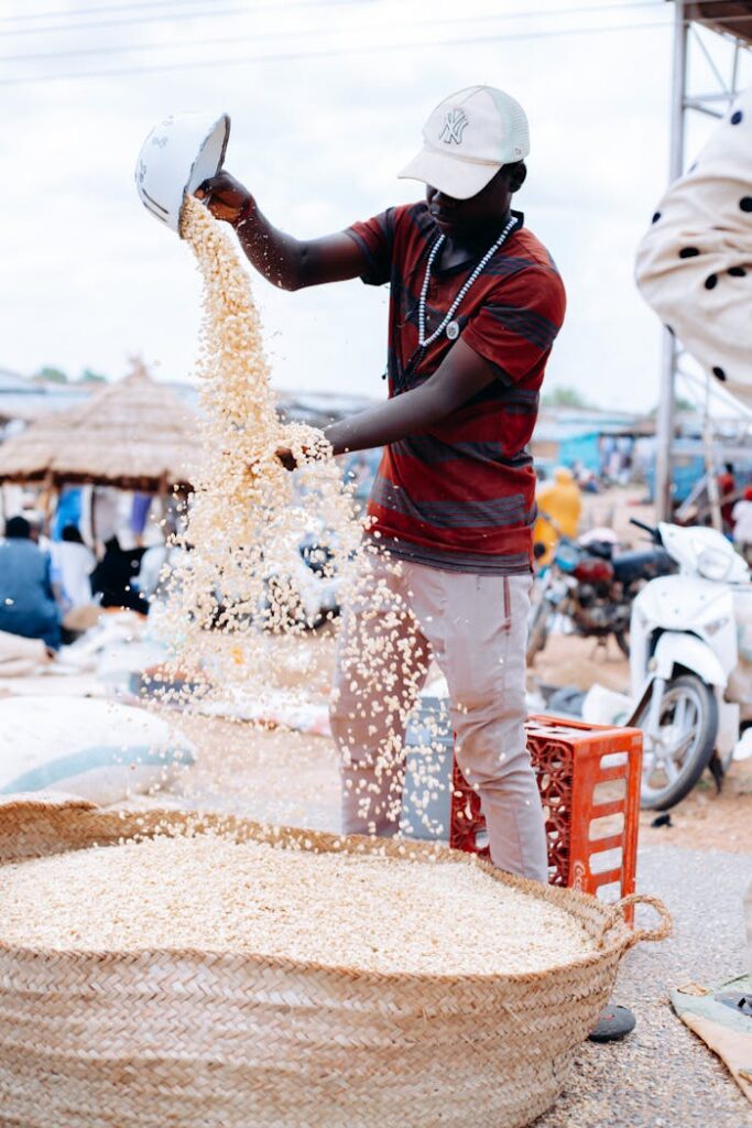 A vendor at an outdoor market pours grains into a large woven basket, symbolizing trade and culture.